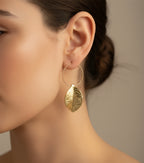 Boucles d’oreilles dorées Sophia en acier inoxydable avec pendentif feuille texturée sur fond marbré clair porté