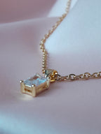 Collier Anastasia doré en acier inoxydable avec pendentif rectangulaire en cristal clair, posé sur tissu rose.