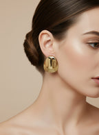 Boucles d’oreilles Daphné dorées en acier inoxydable porté à texture martelée et surface polie, posées sur fond rose satiné avec plume blanche.