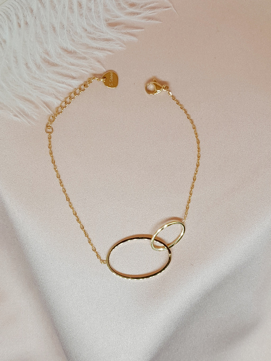 Bracelet Lénae doré à deux anneaux entrelacés en acier inoxydable, taille 16+ cm, posé sur un tissu blanc satiné.