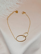 Bracelet Lénae doré à deux anneaux entrelacés en acier inoxydable, taille 16+ cm, posé sur un tissu blanc satiné.