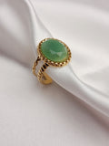 Bague Céleste dorée en acier inoxydable avec pierre verte ovale sur tissu blanc.