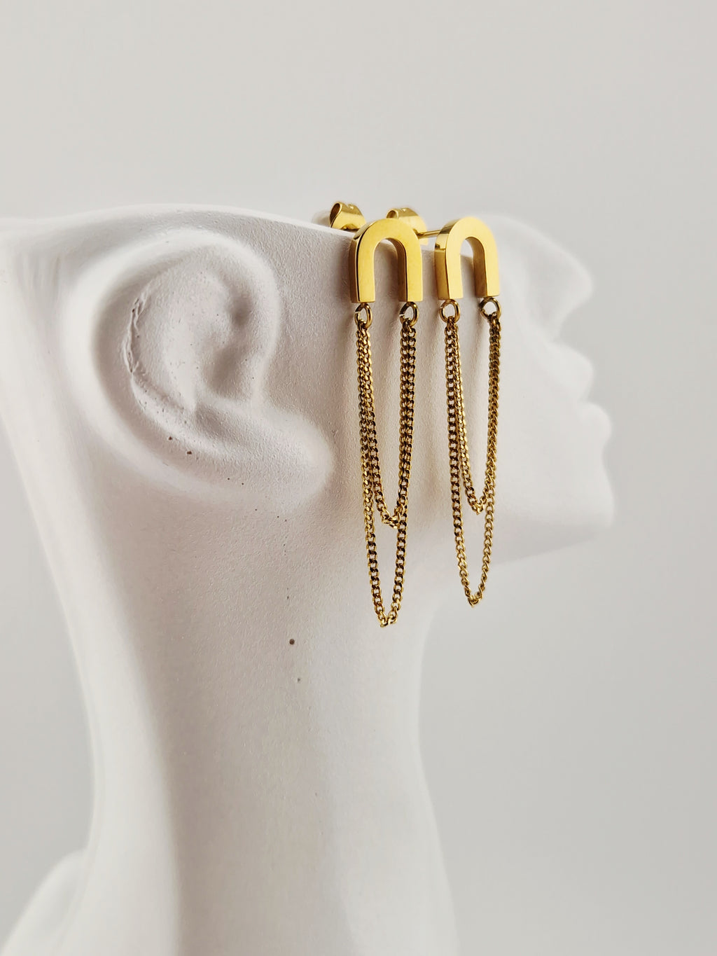 Boucles d’oreilles dorées minimalistes à chaînes pendantes en acier inoxydable, présentées sur un présentoir blanc design.