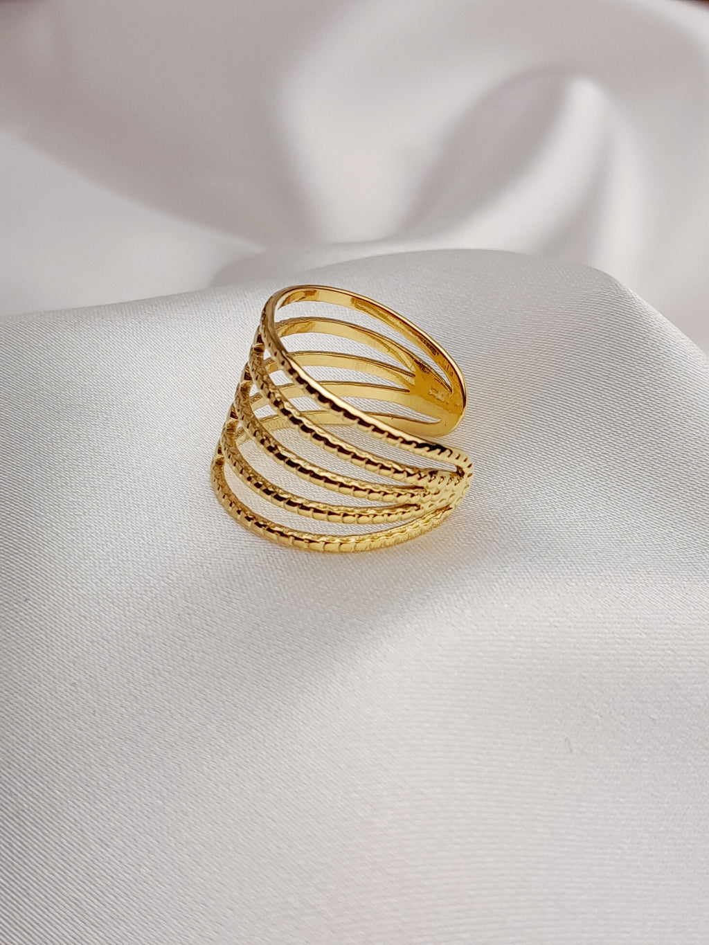 Bague dorée à plusieurs rangs texturés et entrelacés, posée sur un tissu blanc satiné.