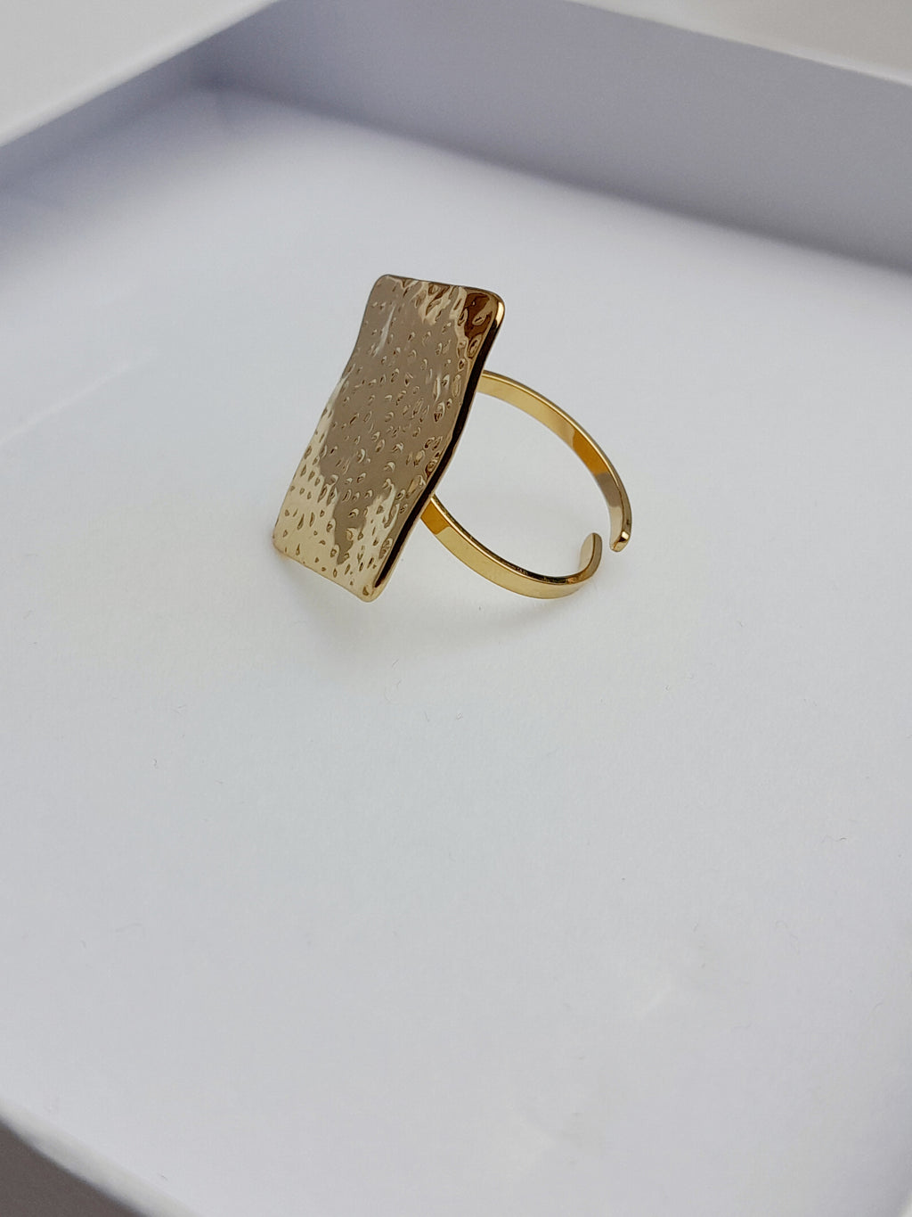 alt="Bague Milla dorée en acier inoxydable avec plateau carré texturé, posée sur tissu satiné blanc – bijou graphique et moderne"
