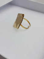 alt="Bague Milla dorée en acier inoxydable avec plateau carré texturé, posée sur tissu satiné blanc – bijou graphique et moderne"
