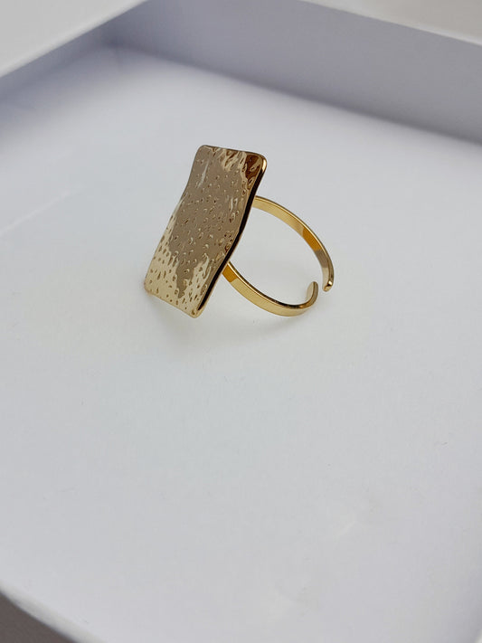 alt="Bague Milla dorée en acier inoxydable avec plateau carré texturé, posée sur tissu satiné blanc – bijou graphique et moderne"
