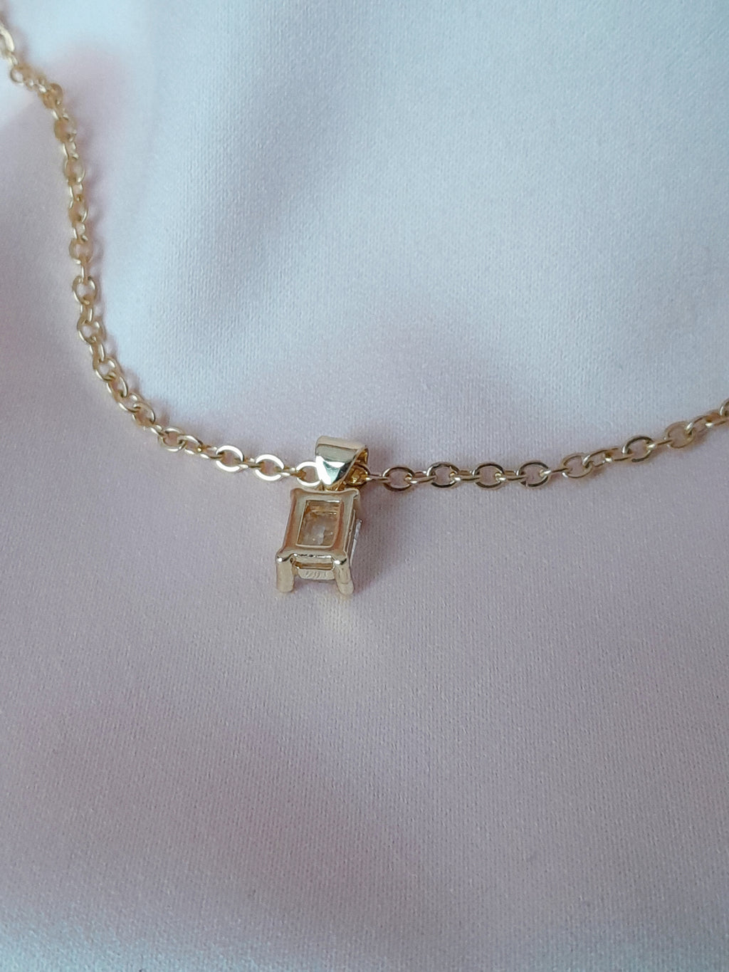 Collier Anastasia doré en acier inoxydable avec pendentif rectangulaire en cristal clair, posé sur tissu rose.