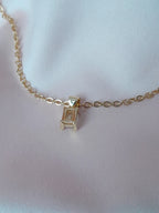 Collier Anastasia doré en acier inoxydable avec pendentif rectangulaire en cristal clair, posé sur tissu rose.