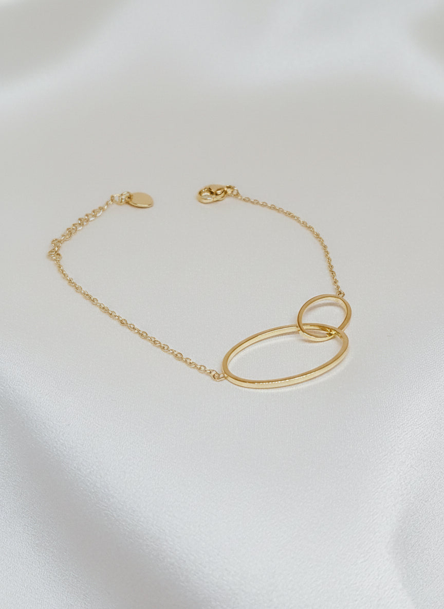 Bracelet Lénae doré à deux anneaux entrelacés en acier inoxydable, taille 16+ cm, posé sur un tissu blanc satiné.