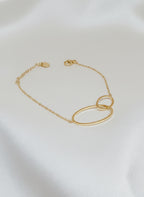 Bracelet Lénae doré à deux anneaux entrelacés en acier inoxydable, taille 16+ cm, posé sur un tissu blanc satiné.