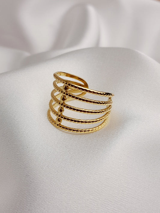 Bague dorée à plusieurs rangs texturés et entrelacés, posée sur un tissu blanc satiné.