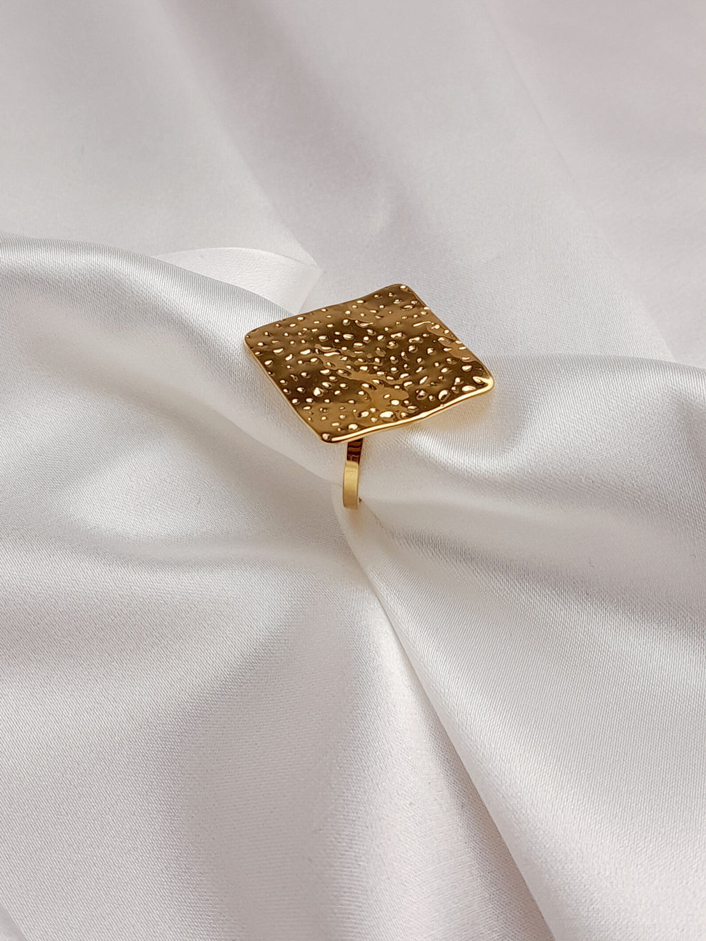 alt="Bague Milla dorée en acier inoxydable avec plateau carré texturé, posée sur tissu satiné blanc – bijou graphique et moderne"
