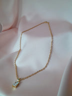 Collier Anastasia doré en acier inoxydable avec pendentif rectangulaire en cristal clair, posé sur tissu rose.