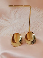 Boucles d’oreilles Daphné dorées en acier inoxydable à texture martelée et surface polie, posées sur fond rose satiné avec plume blanche.