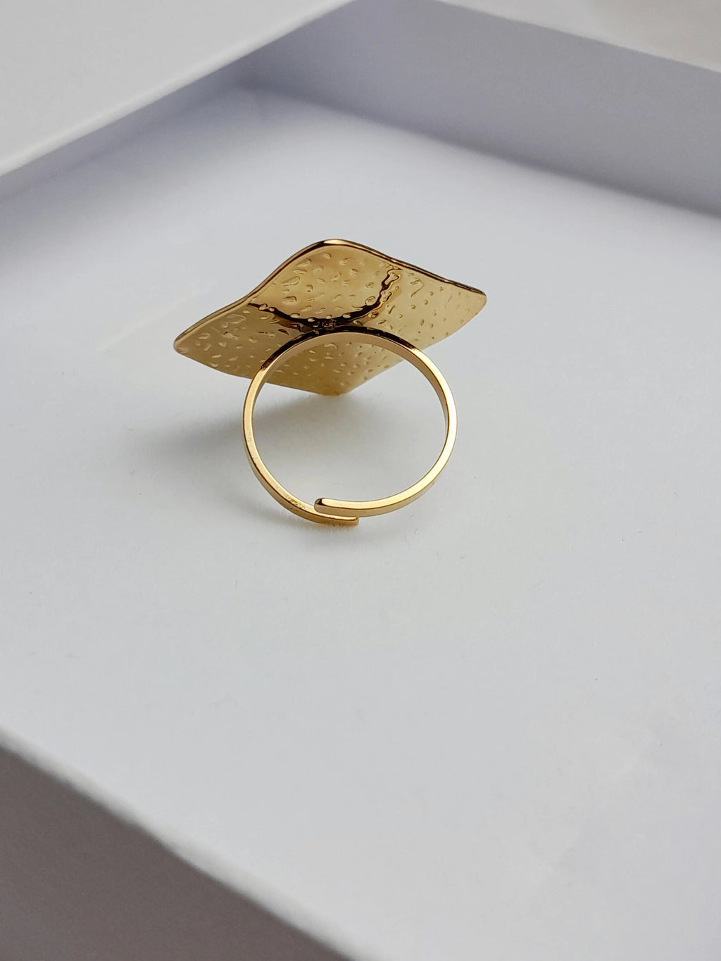 alt="Bague Milla dorée en acier inoxydable avec plateau carré texturé, posée sur tissu satiné blanc – bijou graphique et moderne"
