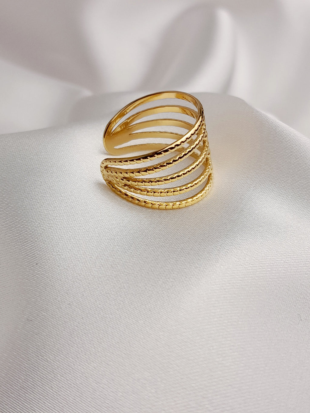 Bague dorée à plusieurs rangs texturés et entrelacés, posée sur un tissu blanc satiné.