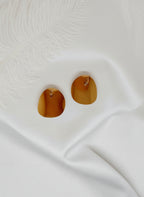 Boucles d’oreilles Daphné dorées en acier inoxydable à texture martelée et surface polie, posées sur fond rose satiné avec plume blanche.