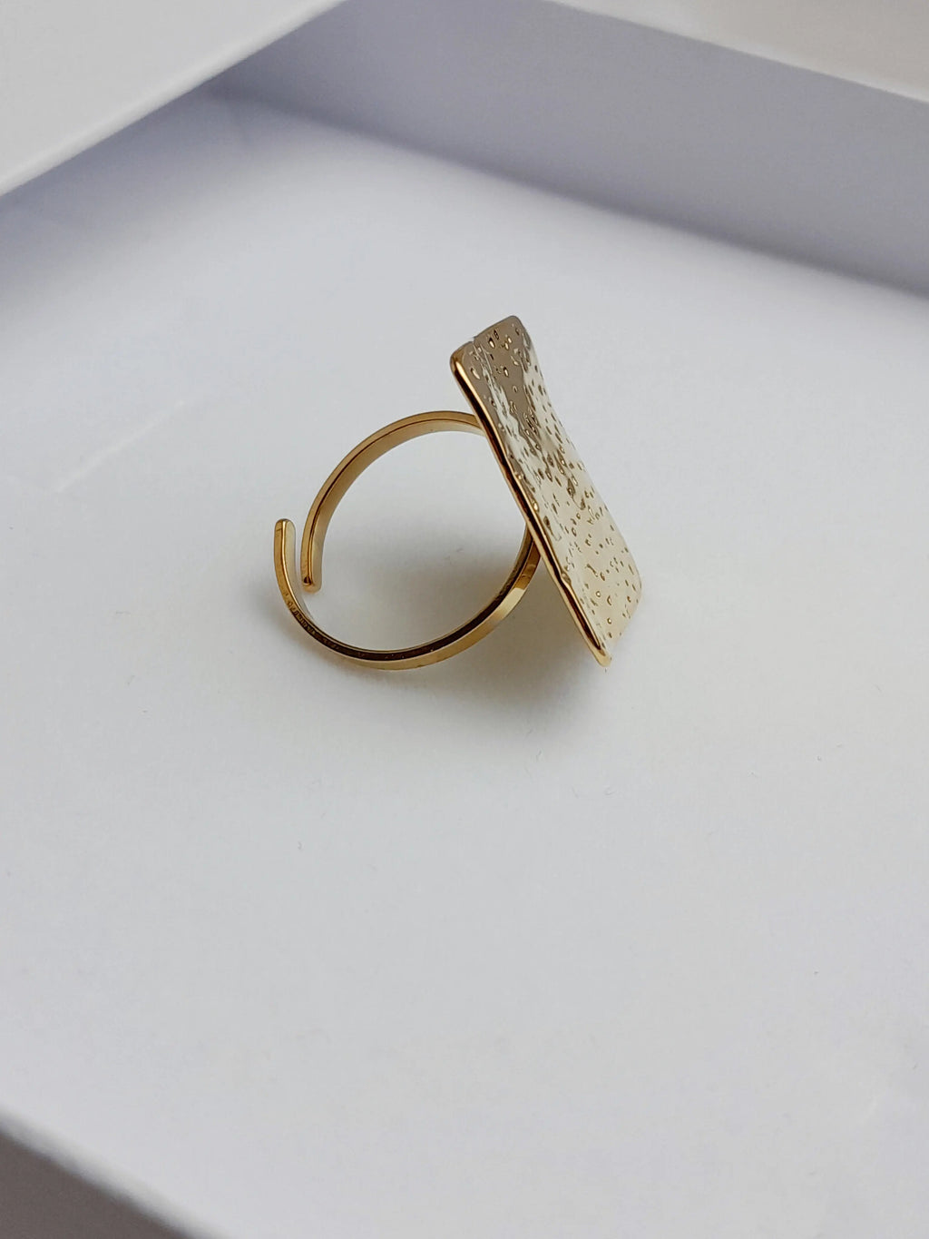 alt="Bague Milla dorée en acier inoxydable avec plateau carré texturé, posée sur tissu satiné blanc – bijou graphique et moderne"
